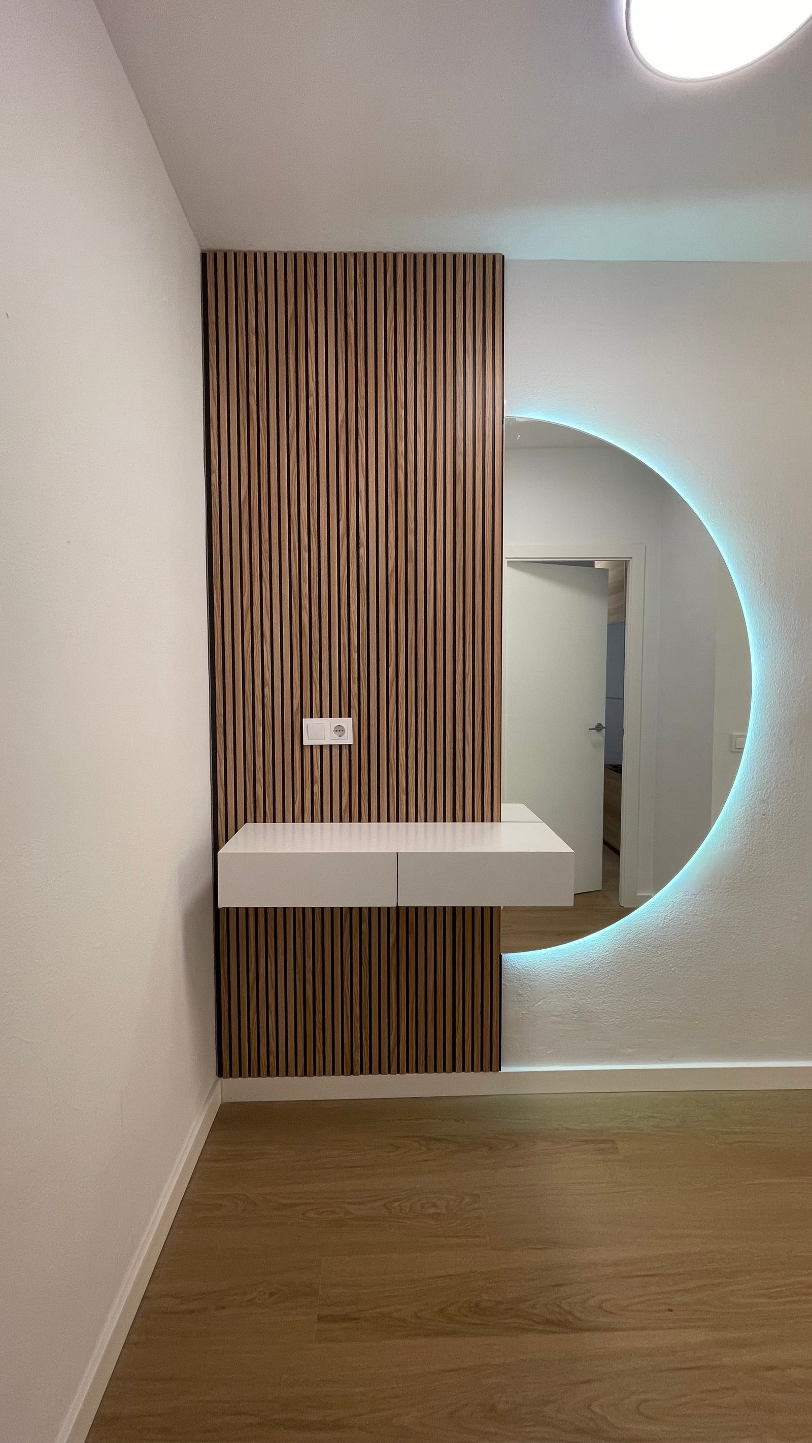 Recibidor con panel de listones de madera, consola flotante y espejo redondo retroiluminado