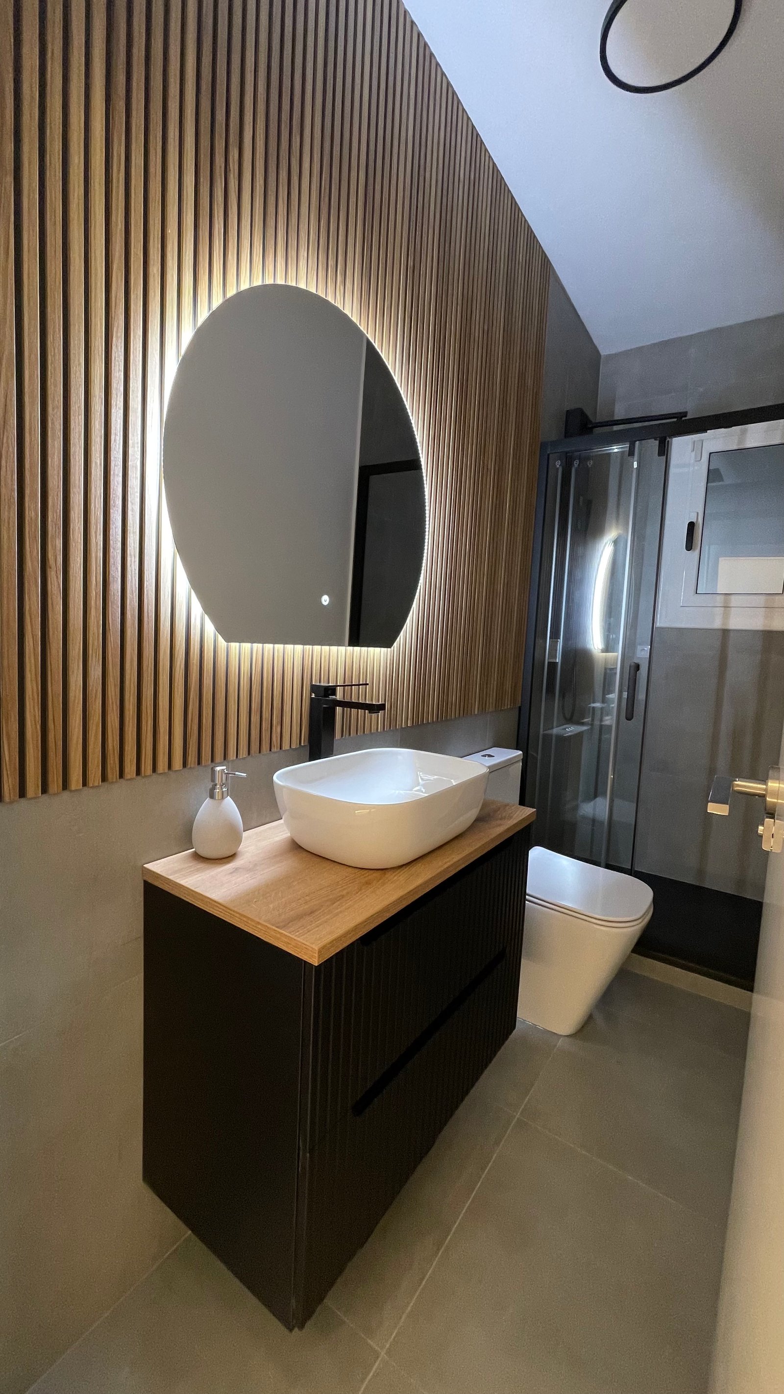 Baño con listones de madera y espejo redondo retroiluminado